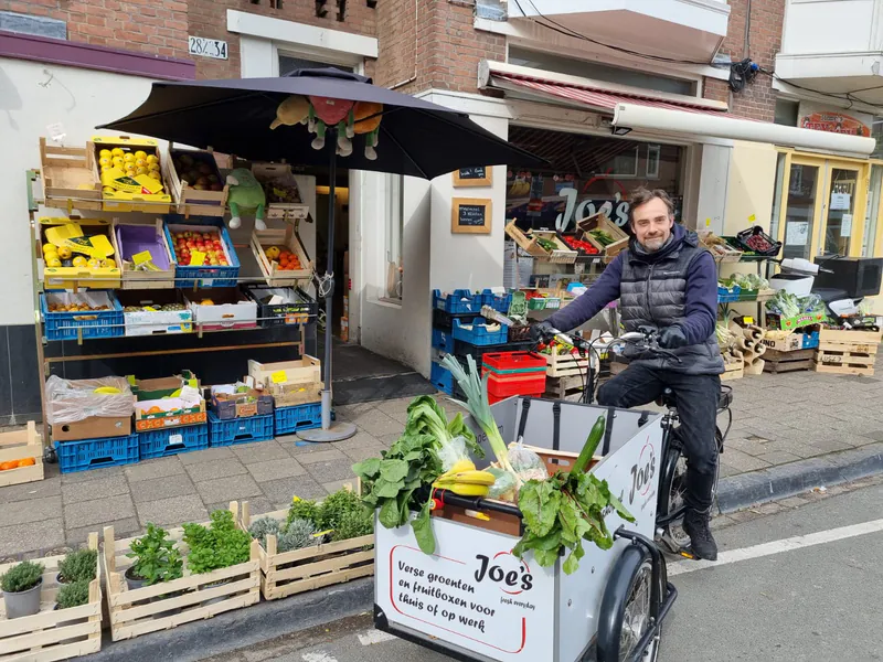 Joe op de bakfiets van Joe's voor de winkel in de Theresiastraat, met kratten groente en fruit op de stoep.