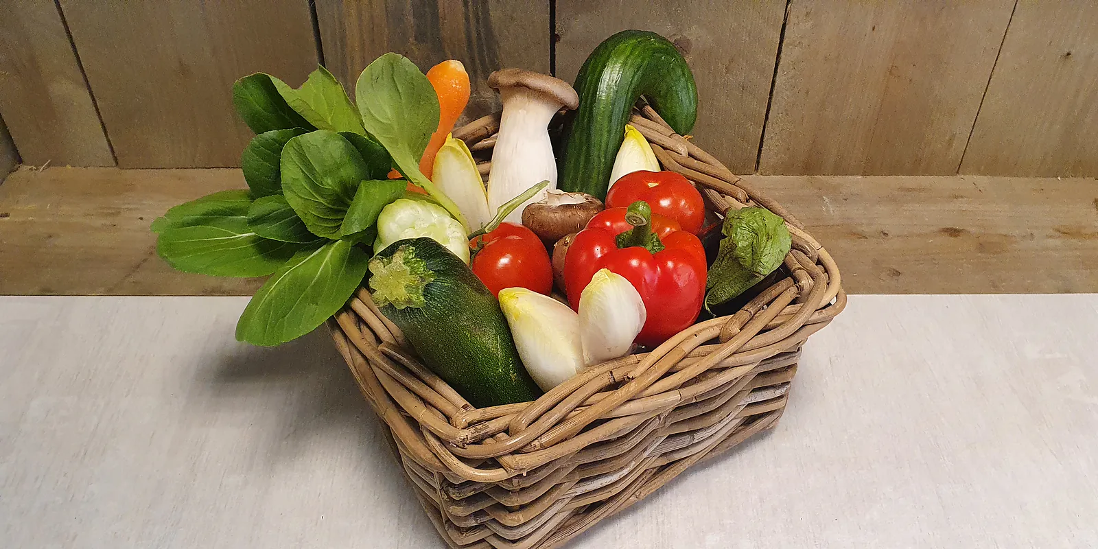 Groentebox klein in een rieten mand op dezelfde plank: paksoi, wortel, courgette, kastanjechampignons, tomaten, paprika en witlof.