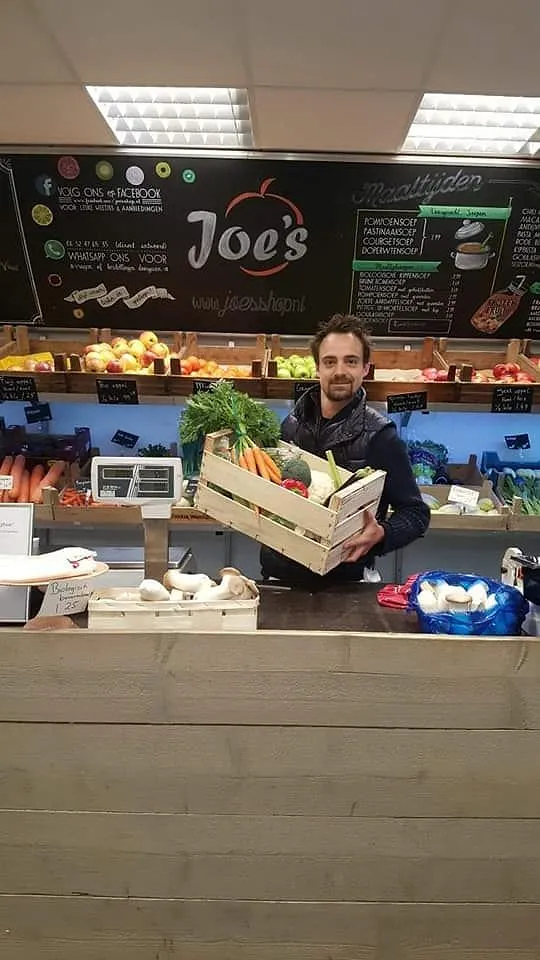 Joe achter de toonbank met een houten kistje groente, onder de krijtborden met het logo.