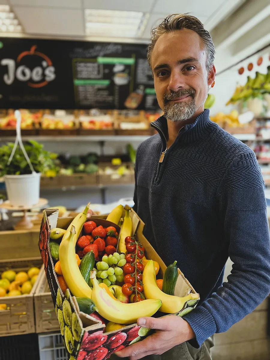 Joe in zijn winkel met een kist vol bananen, aardbeien, druiven en tomaten.