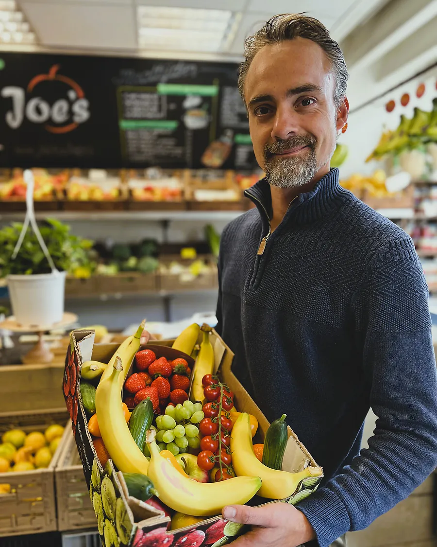 Joe in zijn winkel met een kist vol bananen, aardbeien, druiven en tomaten, achter hem het krijtbord met het logo.